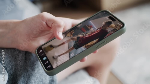 Smartphone application with various video content displayed on phone screen. Close up of teenager scrolling and watching social media feed with short videos using mobile phone while sitting on sofa.