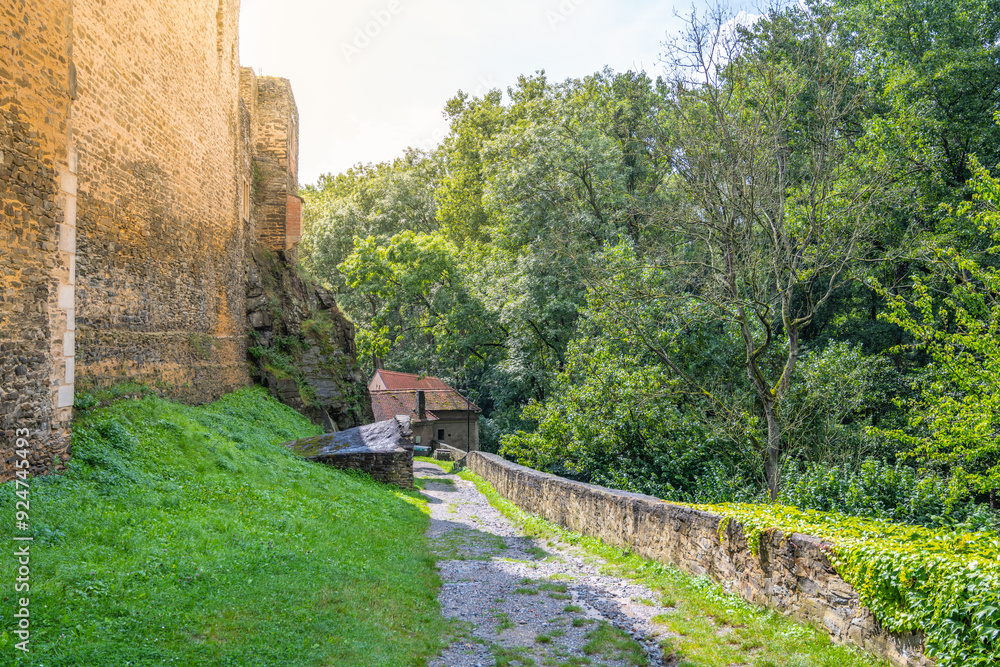 Bright sunlight bathes the lush greenery surrounding the ancient walls of Okor Castle near Prague, with a quiet path inviting exploration. Bright sunlight bathes the lush greenery surrounding the ancient walls of Okor Castle near Prague, with a quiet path inviting exploration.