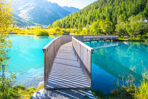 Obraz na plátně A tranquil wooden boardwalk bridge curves above the vibrant blue waters of Lake in Sulden, Italy
