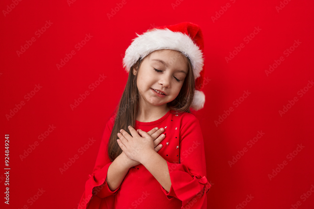 Adorable hispanic girl, happy and smiling, gratefully clasping hands over chest wearing christmas hat. health-proud against isolated red background. Adorable hispanic girl, happy and smiling, gratefully clasping hands over chest wearing christmas hat. health-proud against isolated red background.