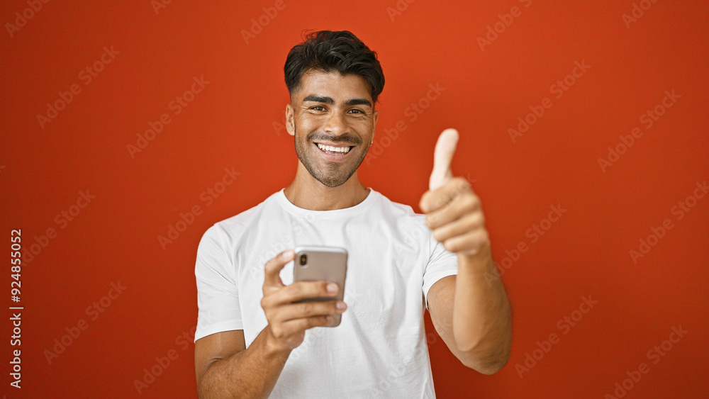 A cheerful young hispanic man with a beard giving a thumbs-up while holding a smartphone against a red wall. A cheerful young hispanic man with a beard giving a thumbs-up while holding a smartphone against a red wall.