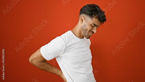 A young hispanic man with a beard winces in pain, touching his lower back against a red wall. photo