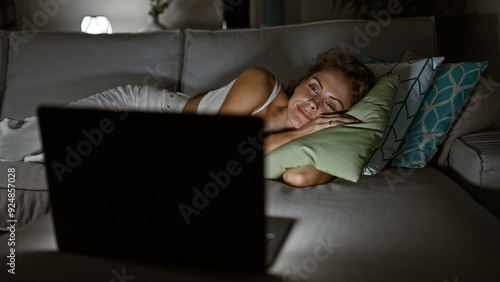 Quadro su tela A young woman relaxes on a couch at home, watching a laptop in a cozy, dimly lit living room