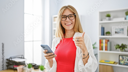 Confident woman in glasses giving thumbs up in bright office, smartphone in hand