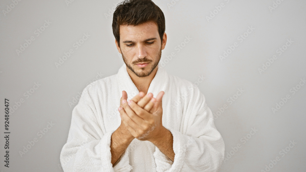 A young hispanic man applies lotion to his hands while wearing a white bathrobe against an isolated background. A young hispanic man applies lotion to his hands while wearing a white bathrobe against an isolated background.