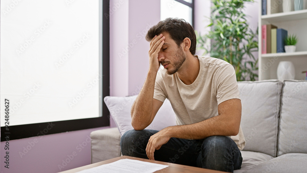A worried young hispanic man with a beard, sitting on a couch indoors, looking at a document. A worried young hispanic man with a beard, sitting on a couch indoors, looking at a document.