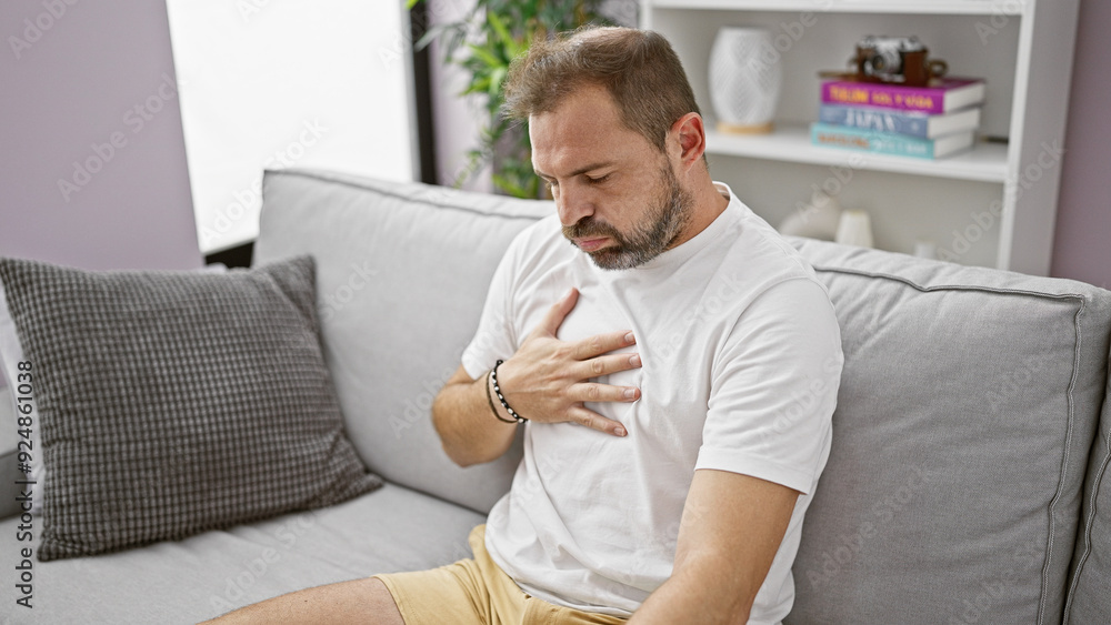 Mature hispanic man with grey hair feeling chest pain in a home interior with sofa background. Mature hispanic man with grey hair feeling chest pain in a home interior with sofa background.