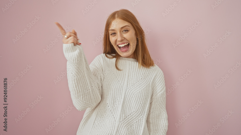 A cheerful young woman with red hair, wearing a sweater, joyfully pointing upwards against a pink background. A cheerful young woman with red hair, wearing a sweater, joyfully pointing upwards against a pink background.