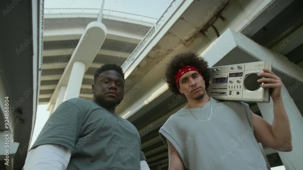 Low angle view of man holding boombox on shoulder and posing for camera with serious expression next to Black friend under urban overpass Low angle view of man holding boombox on shoulder and posing for camera with serious expression next to Black friend under urban overpass