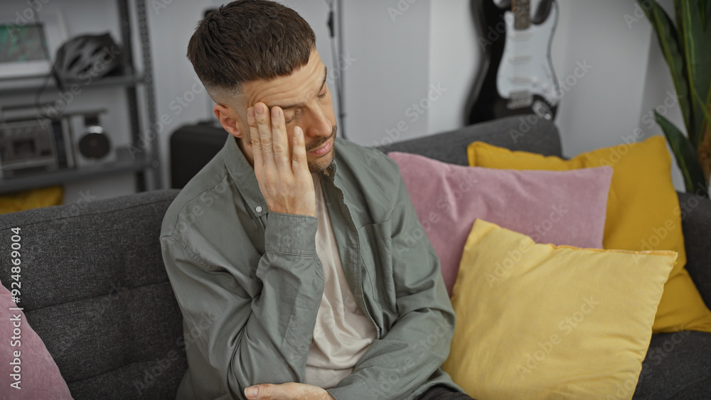 Handsome hispanic man feeling stressed while sitting on a grey sofa in a modern living room setting. Handsome hispanic man feeling stressed while sitting on a grey sofa in a modern living room setting.