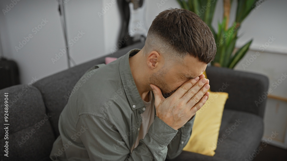 A troubled hispanic man sitting indoors with eyes closed and hands on face, conveying emotion on a couch in a modern living room. A troubled hispanic man sitting indoors with eyes closed and hands on face, conveying emotion on a couch in a modern living room.