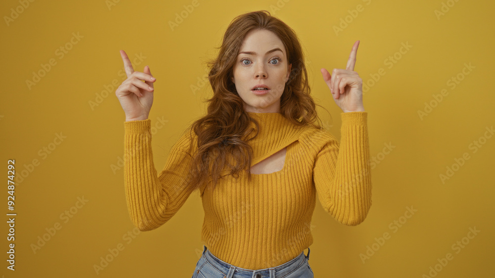 Attractive young caucasian woman with blonde hair and a surprised expression in a yellow sweater pointing upwards, isolated against a yellow background wall. Attractive young caucasian woman with blonde hair and a surprised expression in a yellow sweater pointing upwards, isolated against a yellow background wall.