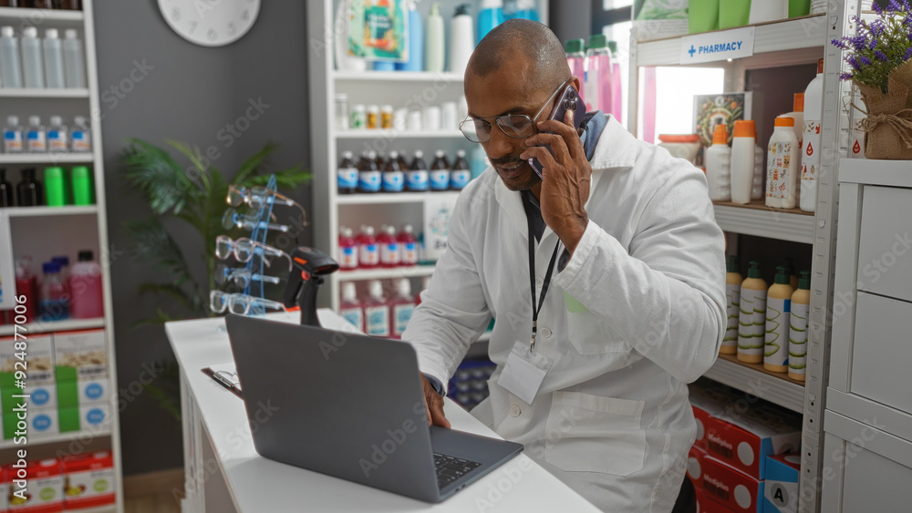 Pharmacist talking on phone while working on laptop in a pharmacy with shelves of products in the background Pharmacist talking on phone while working on laptop in a pharmacy with shelves of products in the background