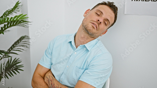 A tired young man with a beard and tattoo sleepily awaits in a clean, minimalist room, signaling a casual break or wait. photo