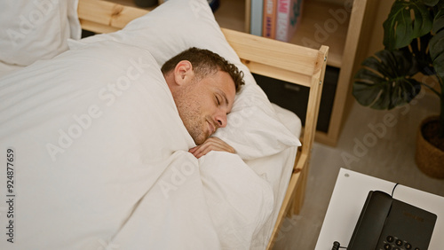 Handsome man sleeping peacefully in a cozy bedroom with soft white bedding and a phone on the nightstand.