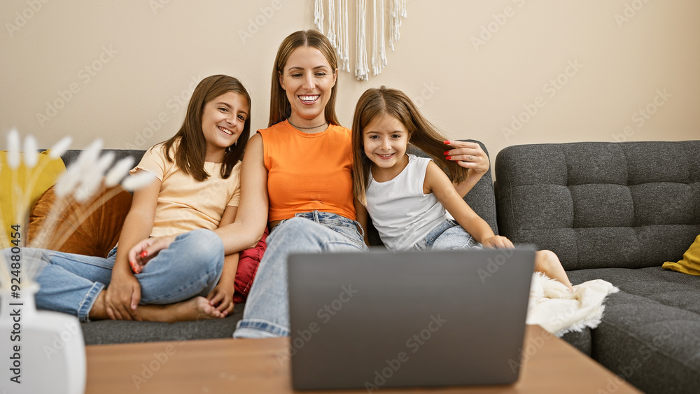 A smiling woman with her two daughters enjoying a moment together indoors, sitting on a gray sofa in front of a laptop. A smiling woman with her two daughters enjoying a moment together indoors, sitting on a gray sofa in front of a laptop.