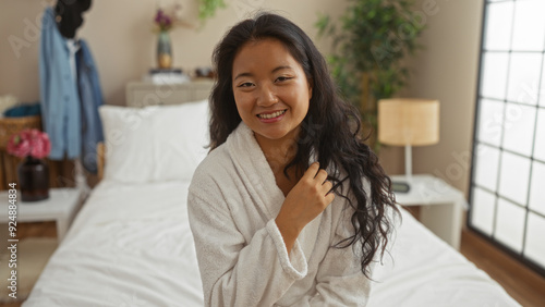 Free Download Beautiful young woman in a bedroom setting, sitting on a bed wearing a white robe, with a happy smile, captured indoors at home, adding a sense of comfort and relaxation. - GFXLoad