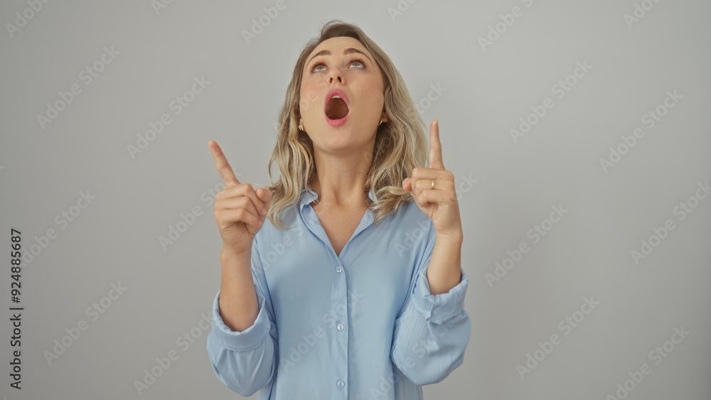 A surprised young woman with blonde hair pointing upwards, isolated against a white background, expressing astonishment. A surprised young woman with blonde hair pointing upwards, isolated against a white background, expressing astonishment.