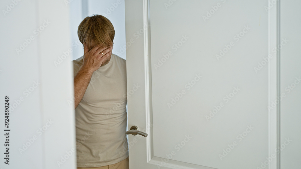 Stressed young adult man with blond beard standing in a home interior, showcasing emotion. Stressed young adult man with blond beard standing in a home interior, showcasing emotion.
