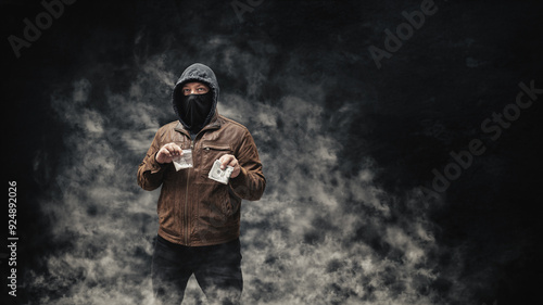 Drug dealer, dangerous hooded criminal man in black mask and brown leather jacket holding transparent plastic bag with drugs and dollar bills, on a dark background with smoke