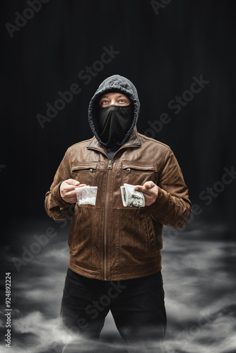 Drug dealer, dangerous hooded criminal man in black mask and brown leather jacket holding transparent plastic bag with drugs and dollar bills, on a dark background with smoke