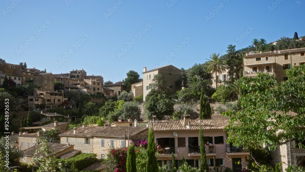 Obraz premium Mallorca village landscape with charming stone houses surrounded by lush greenery and perched on the rolling hills of deia under a clear blue sky.