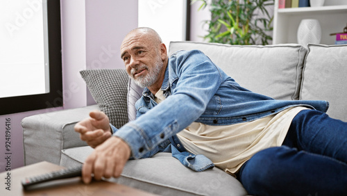 Canvas Print A mature man in casual attire relaxes on a sofa reaching for a remote in a cozy living room