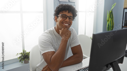 Young man smiling indoors in an office setting while sitting at a desk in front of a computer monitor, exuding a sense of happiness and productivity in a brightly lit room