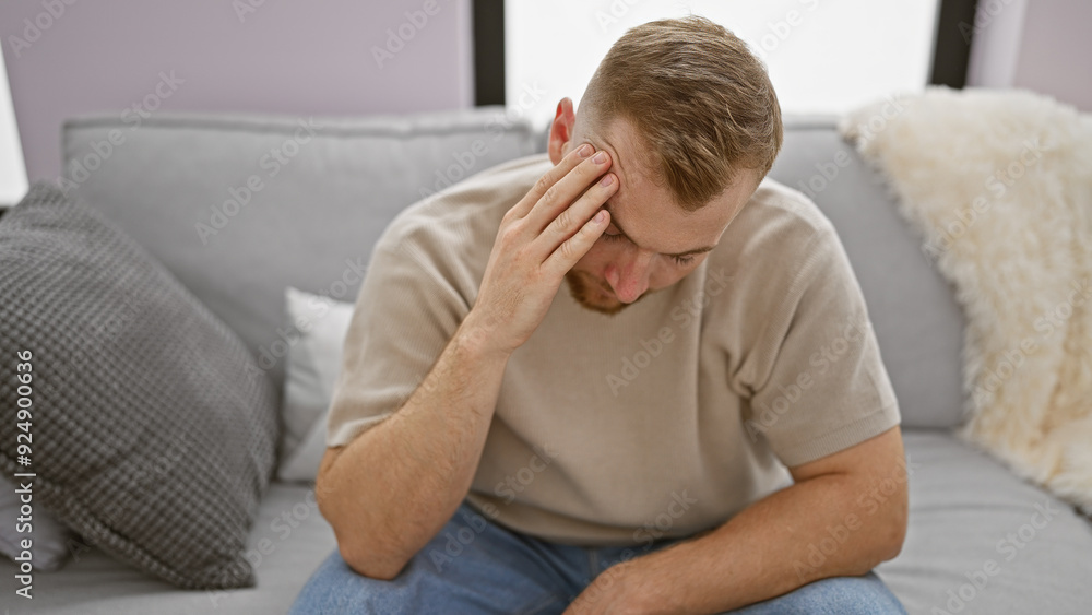 A distressed young caucasian man with a beard sitting on a gray couch indoors appears contemplative or worried. A distressed young caucasian man with a beard sitting on a gray couch indoors appears contemplative or worried.