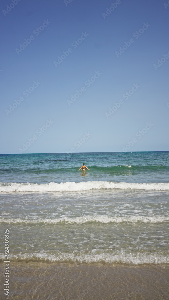 Obraz premium Woman swimming in the ocean with clear blue sky and waves crashing on the sandy shore, evoking a sense of summer and relaxation