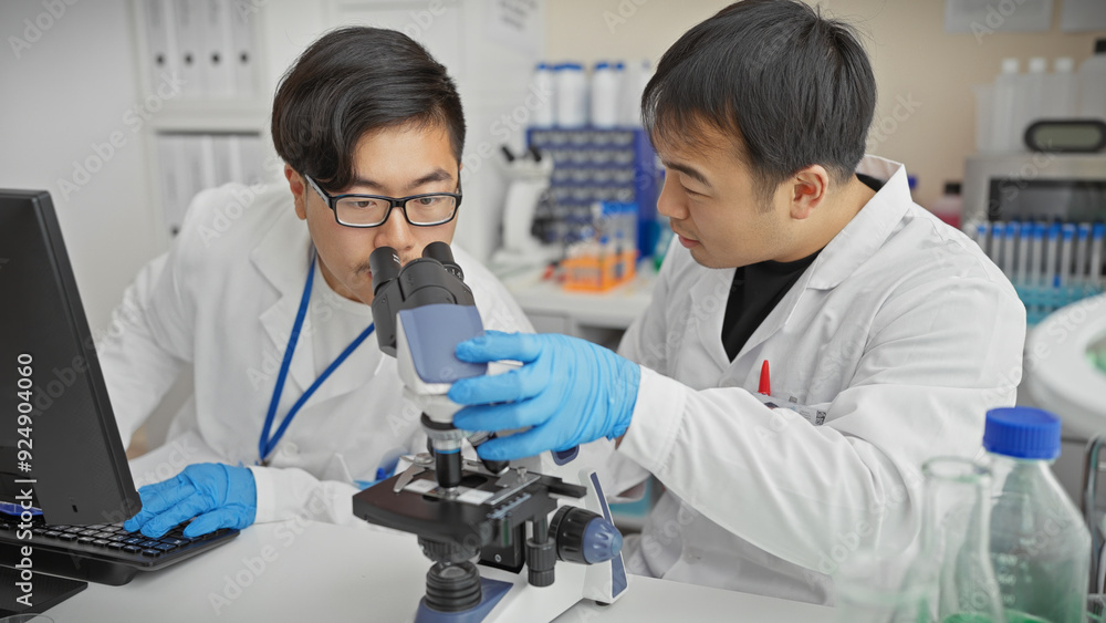 Two men in lab coats work together in a laboratory, one using a microscope, highlighting teamwork in a scientific research setting. Two men in lab coats work together in a laboratory, one using a microscope, highlighting teamwork in a scientific research setting.