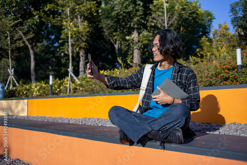 A male student wearing a checked shirt and glasses sits in the park taking a selfie with his phone.