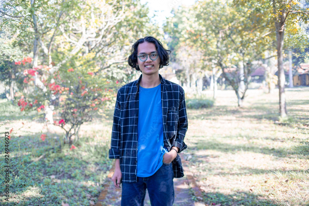 A man wearing a plaid shirt stands in the park casually. A man wearing a plaid shirt stands in the park casually.