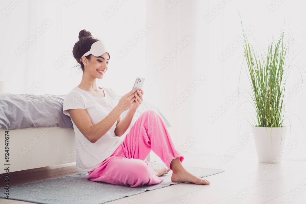 Naklejka premium Photo of beautiful american cute young girl in pajama wake up good morning cozy bedroom indoors with big bed texting using phone sitting floor