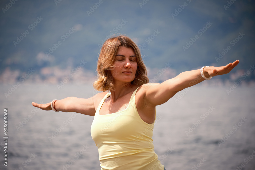 A woman dressed in a yellow top and green leggings is performing a warrior yoga pose by the sea, with majestic mountains and clear sky in the background, promoting strength. A woman dressed in a yellow top and green leggings is performing a warrior yoga pose by the sea, with majestic mountains and clear sky in the background, promoting strength.