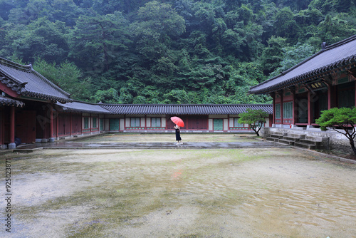 Mungyeongsaejae Pass, Mungyeong-si, Gyeongsangbuk-do, South Korea - July 22, 2022: A female is standing on the yard with red umbrella on a rainy day in froint of Junggungjeon Hall of a movie set photo