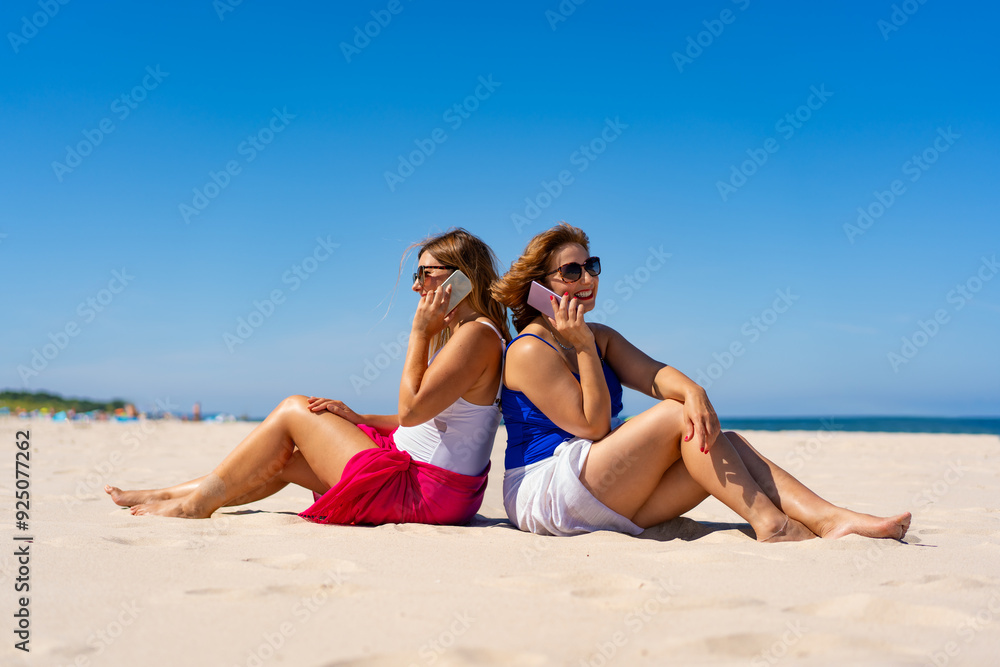Two young beautiful women in fashionable one-piece swimsuits and sarongs and sunglasses sitting on ground leaning back to each other, talking on mobile phones on sandy beach against blue sky in summer