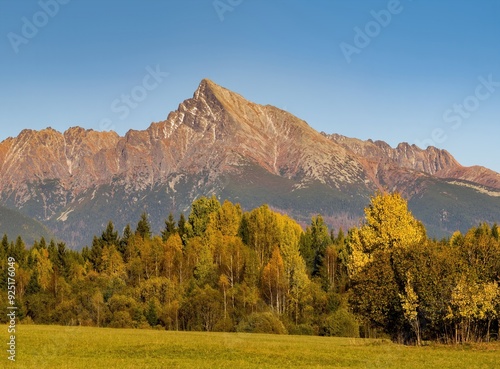 Beautiful autumn alpine landscape with a meadow, colored trees and an old wooden house, in the background a massive mountain ridge. Autumn walks in nature.