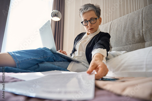 Adult businesswoman in glasses taking documents while working with laptop in hotel room