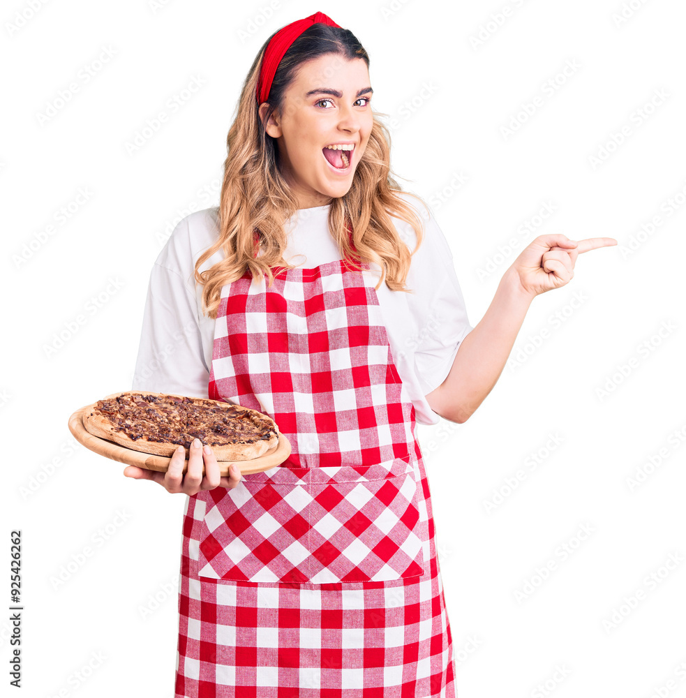 Young caucasian woman wearing apron holding pizza smiling happy pointing with hand and finger to the side