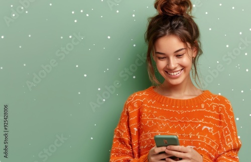 Smiling Woman Holding Smartphone Against Green Wall Wearing Orange Sweater