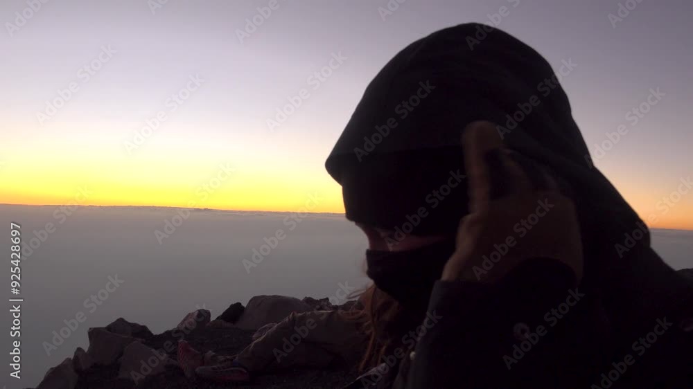 A Woman puts on her neck gaiter and looks at Fuego volcano erupting. Above the clouds at sunset in Guatemala A Woman puts on her neck gaiter and looks at Fuego volcano erupting. Above the clouds at sunset in Guatemala