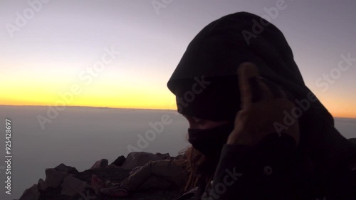 A Woman puts on her neck gaiter and looks at Fuego volcano erupting. Above the clouds at sunset in Guatemala photo