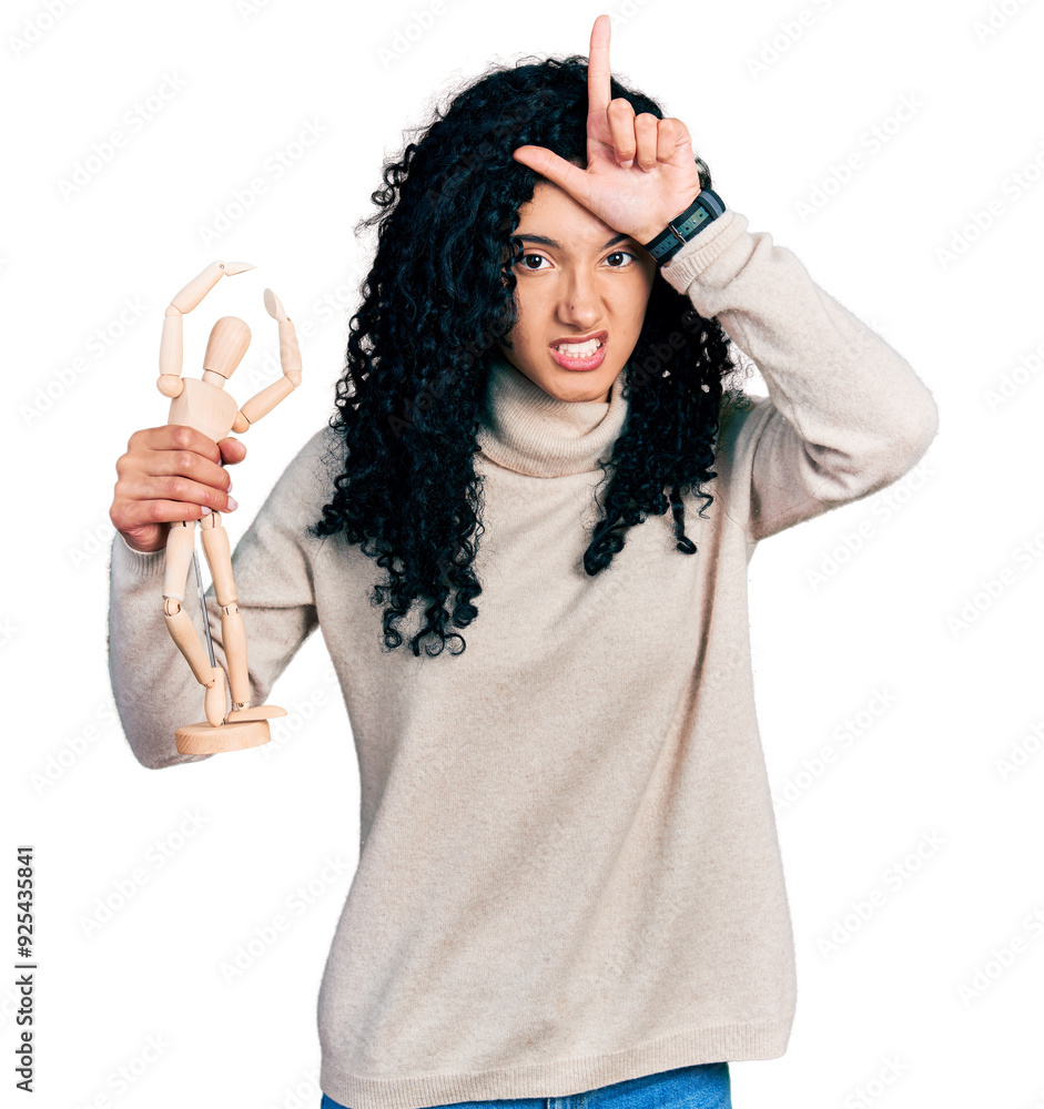 Young hispanic woman with curly hair holding small wooden manikin making fun of people with fingers on forehead doing loser gesture mocking and insulting. Young hispanic woman with curly hair holding small wooden manikin making fun of people with fingers on forehead doing loser gesture mocking and insulting.