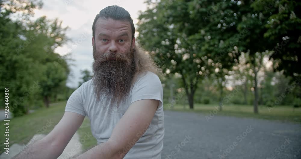 Young confused bearded man in a white t-shirt sits outdoors in a lush green park with trees and a serene atmosphere, looking at the camera. Young confused bearded man in a white t-shirt sits outdoors in a lush green park with trees and a serene atmosphere, looking at the camera.