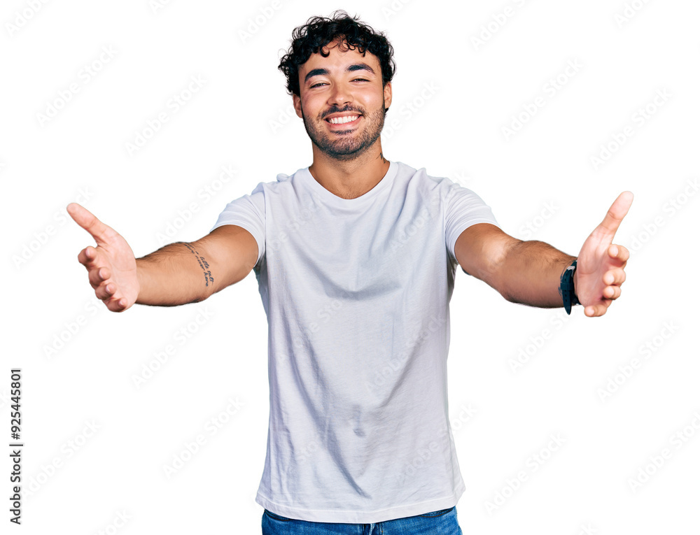 Hispanic young man with beard wearing casual white t shirt looking at the camera smiling with open arms for hug. cheerful expression embracing happiness. Hispanic young man with beard wearing casual white t shirt looking at the camera smiling with open arms for hug. cheerful expression embracing happiness.