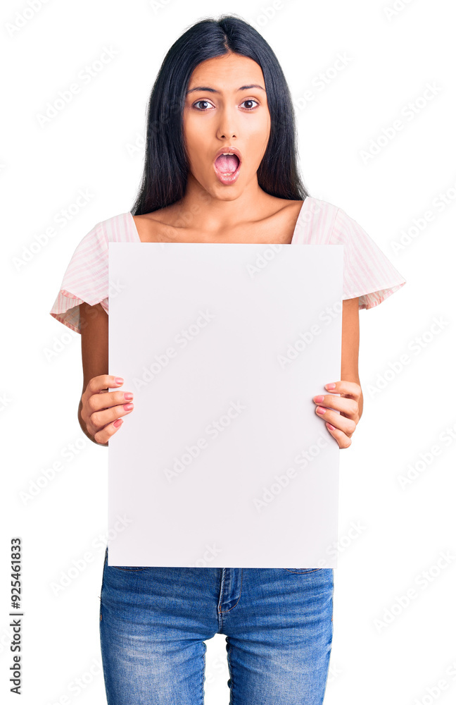 Young beautiful latin girl holding blank empty banner scared and amazed with open mouth for surprise, disbelief face Young beautiful latin girl holding blank empty banner scared and amazed with open mouth for surprise, disbelief face