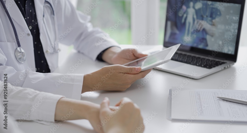 Doctor and a patient. The female physician, wearing a white medical coat over a dark blue dotted blouse, using tablet computer during a consultation in the clinic. Medicine concept Doctor and a patient. The female physician, wearing a white medical coat over a dark blue dotted blouse, using tablet computer during a consultation in the clinic. Medicine concept
