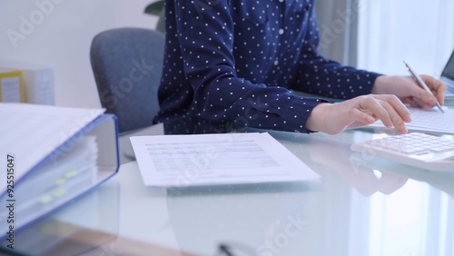 A female accountant with blue dotted blousy is using a laptop computer and calculator to calculate taxes at a glass desk in the office. Right side view photo