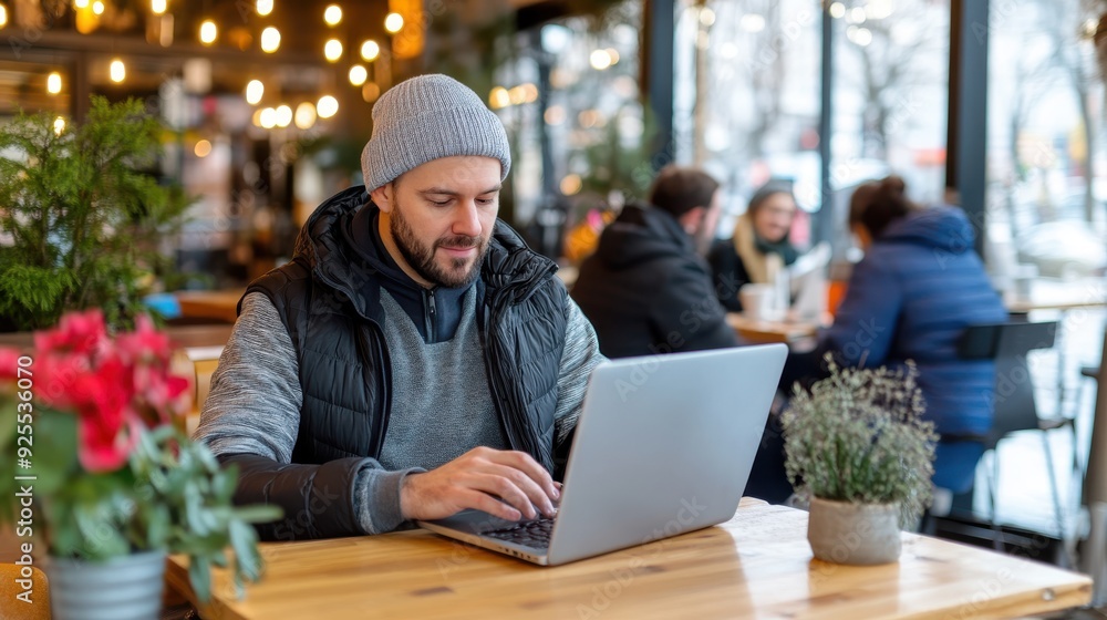 A man wearing a beanie is working on his laptop in a coffee shop, concentrating on his tasks amidst a cozy and warm ambiance with other customers in the background. A man wearing a beanie is working on his laptop in a coffee shop, concentrating on his tasks amidst a cozy and warm ambiance with other customers in the background.